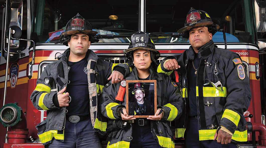 Three firefighters stand before a fire truck as one holds a framed portrait of a fellow firefighter